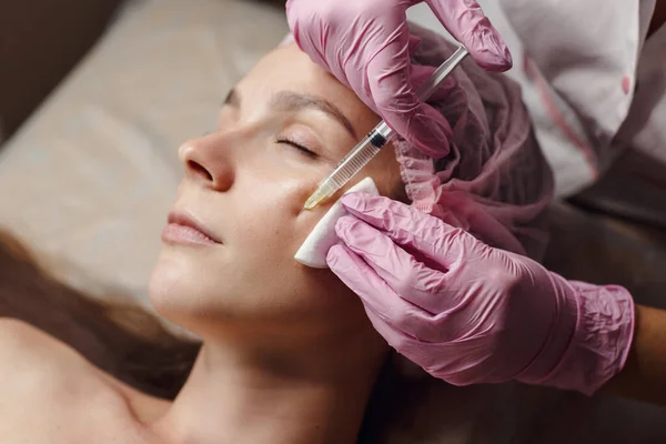 Woman on rejuvenation procedure in beauty clinic filler injection. She lying calmly at clinic. The expert beautician in pink gloves is filling female wrinkles by hyaluronic acid. Injection of beauty.