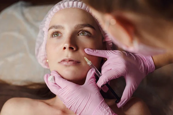 Woman on rejuvenation procedure in beauty clinic filler injection. She lying calmly at clinic. The expert beautician in pink gloves is filling female wrinkles by hyaluronic acid. Injection of beauty.