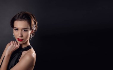 Zarif dalgalı saç stili, parlak makyajlı güzel bir kadın. Siyah elbise giyen, siyah arka planda yalnız poz veren moda esmeri. Göz kapağı oklarında ve kırmızı rujda klasik makyaj.