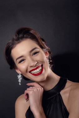 Zarif dalgalı saç stili, parlak makyajlı güzel bir kadın. Siyah elbise giyen, siyah arka planda yalnız poz veren moda esmeri. Göz kapağı oklarında ve kırmızı rujda klasik makyaj.