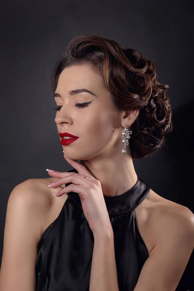 Beautiful gorgeous woman with elegant wavy hairstyle, bright make up. Fashion brunette wearing black dress posing isolated on black background. Classic makeup in eyelid arrows and red lipstick