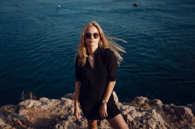 Siyah elbiseli kadın yaz denizinin üzerinde uçurumun kenarında tek başına duruyor. Seyahat yaşam tarzı konsepti.