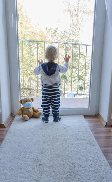 Child in home quarantine playing at the window with his sick teddy bear wearing a medical mask against viruses during coronavirus and flu outbreak. Children and illness COVID-2019 disease concept