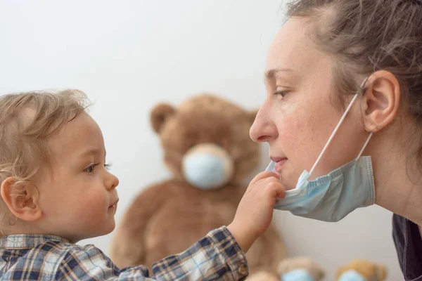 Mother teaching her child about medical mask protection against viruses, by playing with his sick teddy bears. Children and flu or coronavirus COVID-19 illness concept. Selective focus
