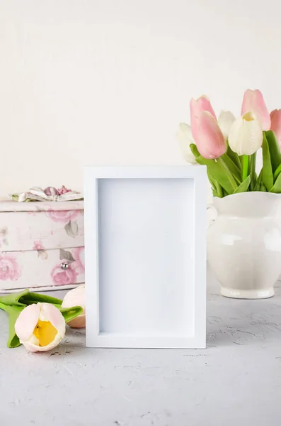Pastel tulip bouquet in ceramic pitcher with empty white frame on shelf. Spring or holiday concept, Easter or Valentine\'s day, birthday. Copy space, template mockup greeting card,  place for text,