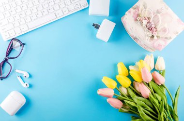 Beyaz klavyenin üst görüntüsü, gözlükler, kulaklıklar, kozmetik ürünleri ve mavi üzerine çiçekler. Serbest çalışan bir kadın. Ofis blogcusu çalışma alanını kavra. Post sosyal medya için şablon düzenleme modellemesi