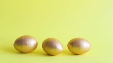 Altın dekoratif yumurtalar yuvarlanıyor ve birbirlerine sarı bir zemin vuruyorlar. Bahar tatili konsepti. Boşluğu kopyala, şablon modelleme Paskalya tebrik kartı 