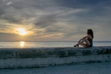 Günbatımına bakan genç bir kadın Küba, Havana 'da iskelede oturuyor.