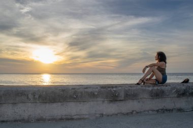 Günbatımına bakan genç bir kadın Küba, Havana 'da iskelede oturuyor.