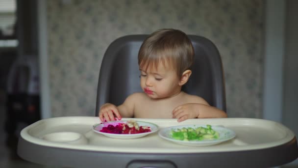 Девочка ест фрукты и овощи, сидя в белом стульчике — стоковое видео