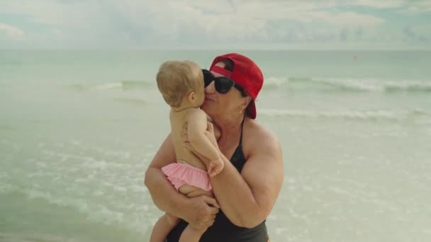 Glückliche Großmutter mit ihrem kleinen Enkelkind am Strand mit dem Meer im Hintergrund — Stockvideo