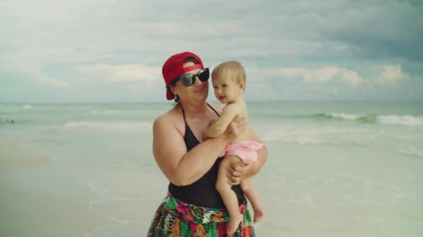 Mutlu büyükanne, arka planda okyanus olan küçük torununu kumsalda tutuyor. — Stok video