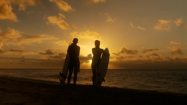 Pareja de surfistas en silueta sosteniendo largas tablas de surf al atardecer en la playa tropical — Vídeos de Stock
