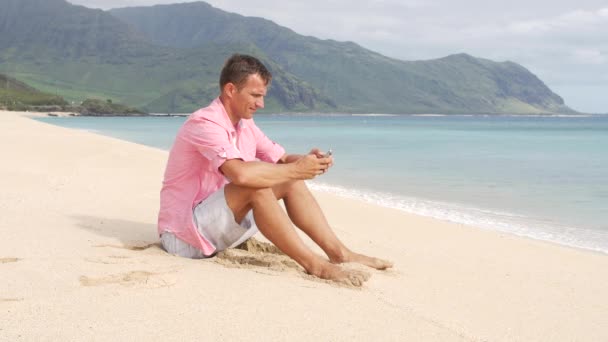Человек со смартфоном сидит на пляже — стоковое видео