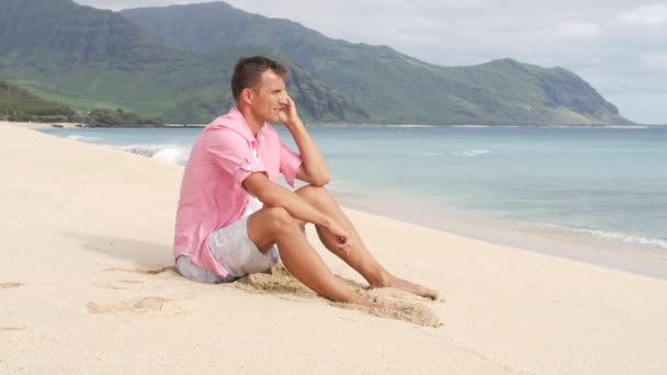 Человек со смартфоном сидит и разговаривает на пляже — стоковое видео