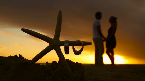 Anillos de boda en la estrella de mar y pareja besándose al atardecer playa amor concepto — Vídeos de Stock