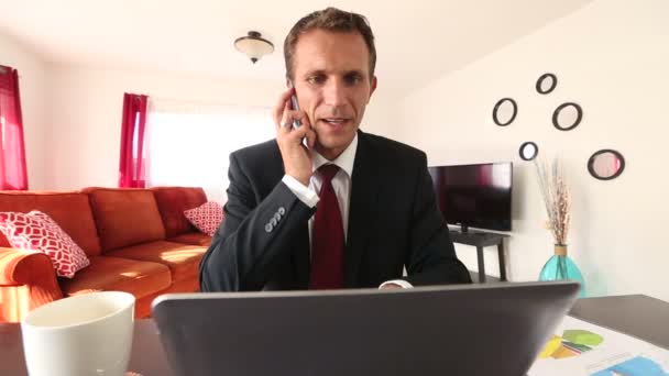 Кавказький чоловік працює на ноутбук і говорити на телефон вдома — стокове відео