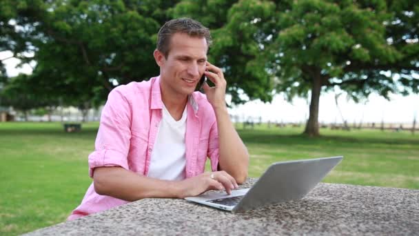 Hombre con el ordenador portátil hablando en el teléfono móvil en el parque — Vídeo de stock