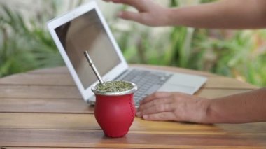 Dizüstü bilgisayarda çalışan ve ev ofisinde çay içen bir kadın. Dişi eller dizüstü bilgisayarda yazı yazıyor, sıcak yerba mate çayı içiyor. Yavaş Hareket.