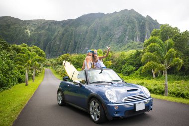 Araba yolculuğu tatilinde gençler yaz tatili sırasında telefonlarıyla selfie çekiyorlar. Turistler, Hawaii 'de üstü açık arabalarla fotoğraf çekiyorlar, akıllı telefon kameralarıyla.