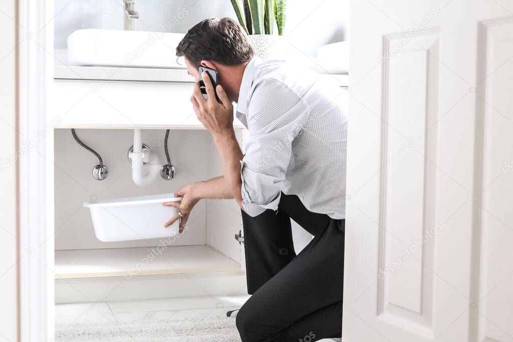 Sad Young Man Calling Plumber In Front Of Water Leaking From Sink Pipe