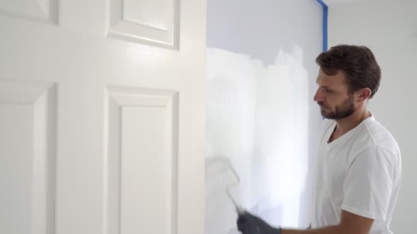 画家の男性は家の中で壁をペイントローラーと白の色の塗料で塗装します 家の部屋の改装 — ストック動画