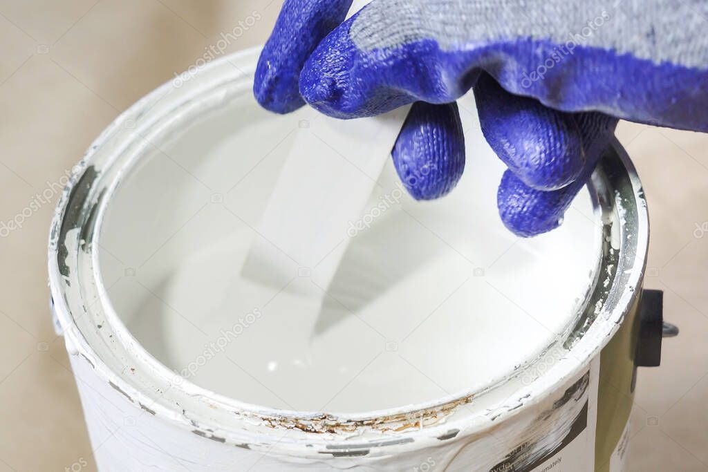 Close up of hand gloved painter mixing paint in metal paint can with wooden stick.