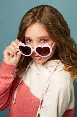 Stüdyoda mavi arka planda poz veren esmer kız. Elma şeklinde pembe gözlüklü kız..