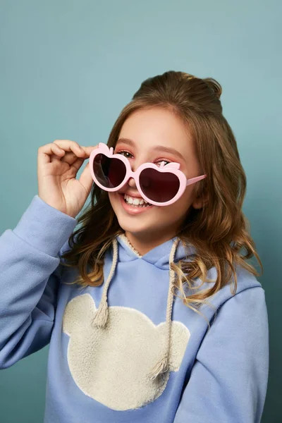 Chica morena glamurosa posando sobre un fondo azul, sosteniendo gafas con estilo con su mano . — Foto de Stock