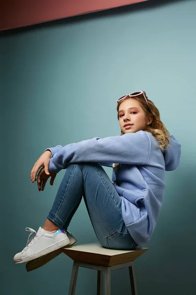 Стильная юная брюнетка в молодежной одежде сидит на стуле с согнутыми ногами. Фотосессия на синем фоне — стоковое фото