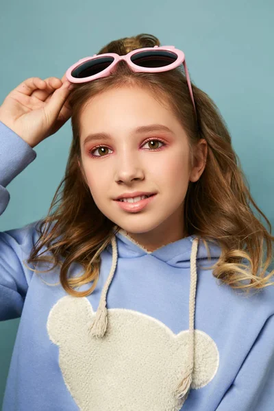 Linda chica en un suéter cálido y gafas de color rosa posando en el estudio sobre un fondo azul. Morena en un suéter caliente con una silueta de oso . — Foto de Stock