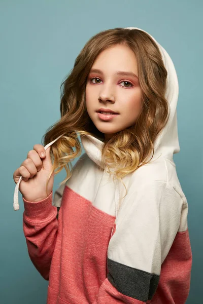 Menina bonito em um suéter multi-colorido com um capuz posando em um fundo azul no estúdio. Morena com maquiagem brilhante segurando uma corda de uma camisola com a mão . — Fotografia de Stock