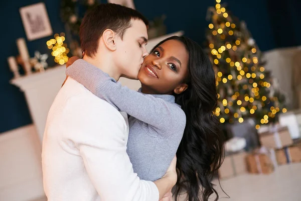 Beautiful interracial young couple celebrating Christmas at home. Christmas tree decorated with a garland. A white guy hugs and kisses a black girl. — 스톡 사진