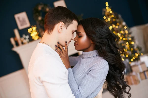 Carino interrazziale coppia sposata in una serata di Natale festiva. Interno decorato per il nuovo anno. nero ragazza abbraccia un bianco ragazzo con la sua occhi chiusi . Immagine Stock