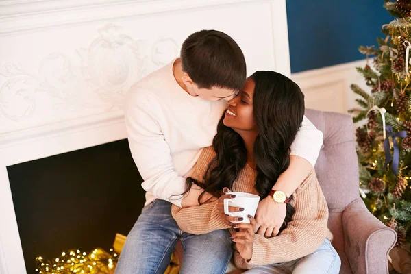 クリスマスの休日の朝に居心地の良い家で若い異人種間のカップル。黒の女の子がお茶を飲んでいる。白人男抱擁A女の子座っていますオンザカウチ. ロイヤリティフリーのストック画像