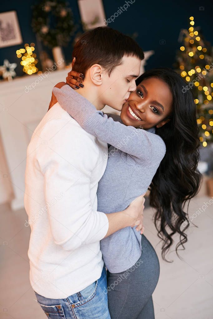 Beautiful black girl and white guy cuddling in the living room on Christmas night. Interracial Relations. In love newlyweds. New Year celebration.