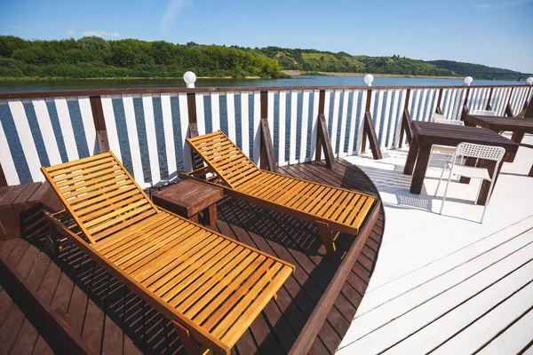 Wooden deck chairs on the landing stage. A place of rest in the open air. Stylish wooden tables. View of the river and green trees — 스톡 사진