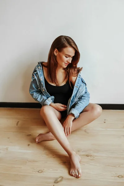 Elegante ragazza durante la gravidanza seduta su un pavimento di legno in posa in una giacca di jeans. Donna felice tiene pancia con mano — Foto Stock