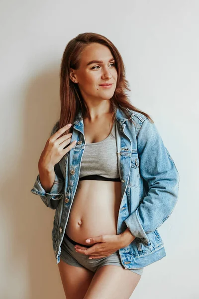 Mooi, stijlvol meisje in een spijkerjasje tijdens de zwangerschap. Vrouw glimlachende hand met afgeronde buik. — Stockfoto