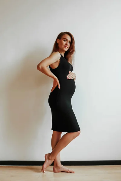 Mooi zwanger meisje in een klassieke zwarte jurk op een achtergrond van een witte muur. Jonge moeder in afwachting van de geboorte van een kind. — Stockfoto