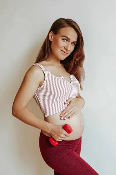 Menina bonita nova fazendo fitness em casa. Aumenta o peso com a mão enquanto segura o estômago. Mulher feliz antes do parto . — Fotografia de Stock