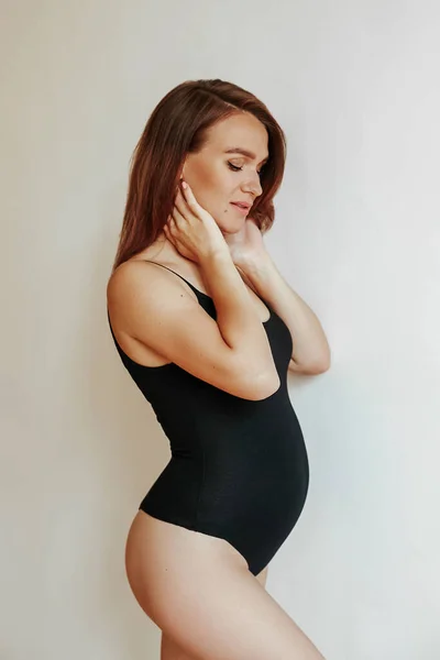 Menina grávida bonito em elegante bodysuit preto de pé lateralmente posando em estúdio. Jovem mãe antes do parto. Menina em posição . — Fotografia de Stock