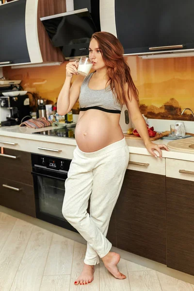 Uma jovem mãe grávida bebe leite enquanto está na cozinha, apoiada na bancada. Menina com cabelo luxuoso e uma barriga grande . — Fotografia de Stock