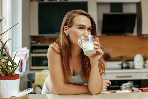 Těhotná dívka v kuchyni drží v ruce sklenici mléka. Mladá matka se opřela o kuchyňský stůl — Stock fotografie