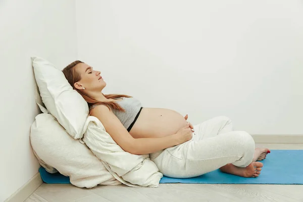 Durante a gravidez, uma jovem menina bonita está apoiada em travesseiros macios e segura a barriga com as mãos. Uma mulher está descansando antes do parto . — Fotografia de Stock