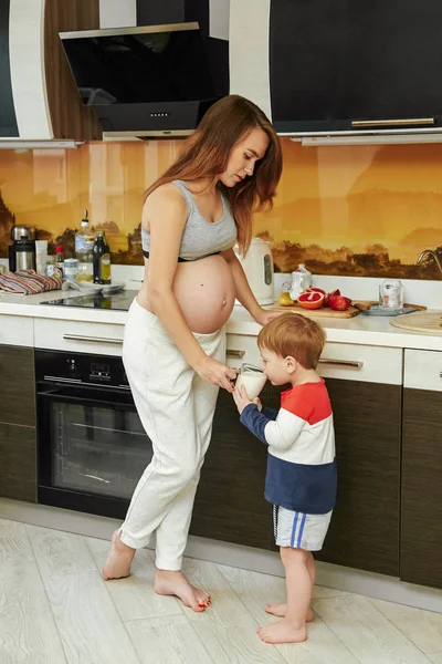 A young pregnant mom gives her little son a mug of milk. A child drinks milk from a glass mug in the kitchen. Breakfast.