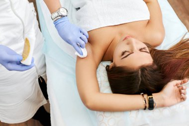 Güzellik salonunda gösterişli bir esmer. Bir kozmetolog kızın vücudunda tüy yolma prosedürü uygular. Şekerleme.