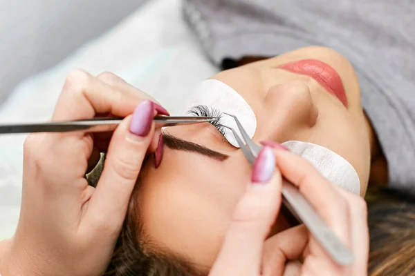 Beautician works with tweezers on the procedure for eyelash extension of a young, beautiful brunette. Beauty saloon.