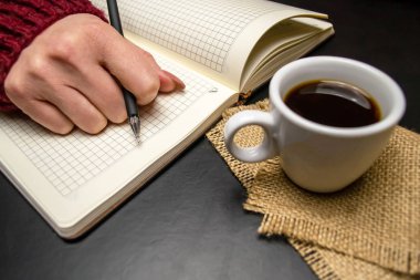 Tükenmez kalemden bir el siyah arka planda bir fincan kahvenin yanındaki deftere uzanır.