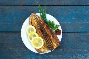Mavi ahşap bir masada beyaz bir tabakta yeşil ve limon dilimli fırında uskumru balığı.